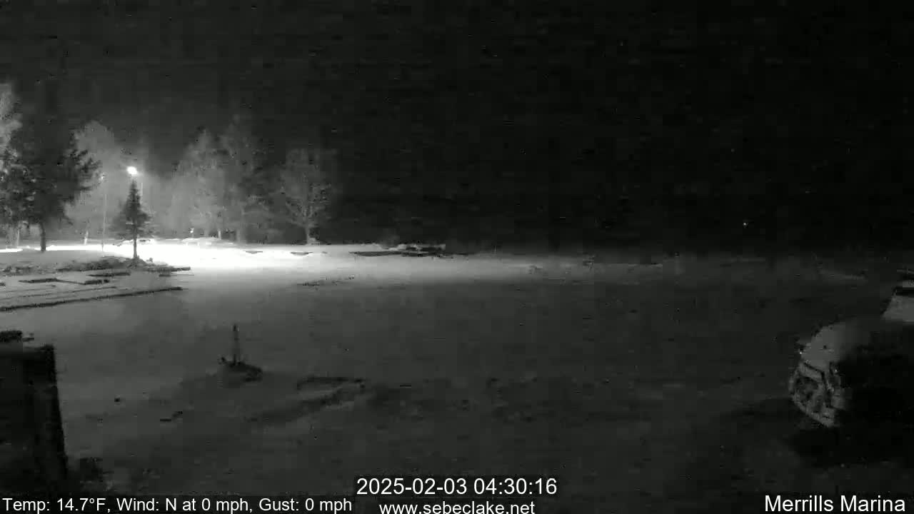 A snow-covered nighttime parking lot is dimly lit, with a few trees and a vehicle visible in the distance under a dark sky.