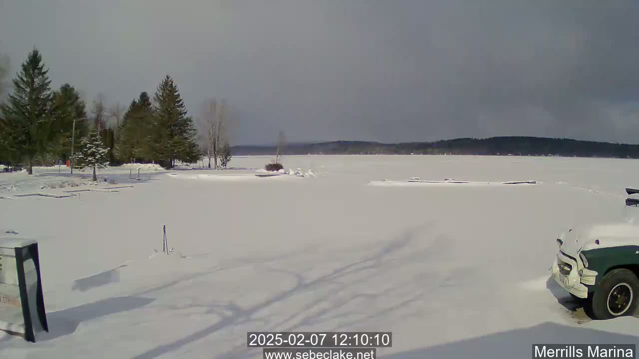 A snow-covered shoreline with evergreen trees and a partially visible truck in the lower right corner, overlooking a frozen lake under a mostly cloudy sky.