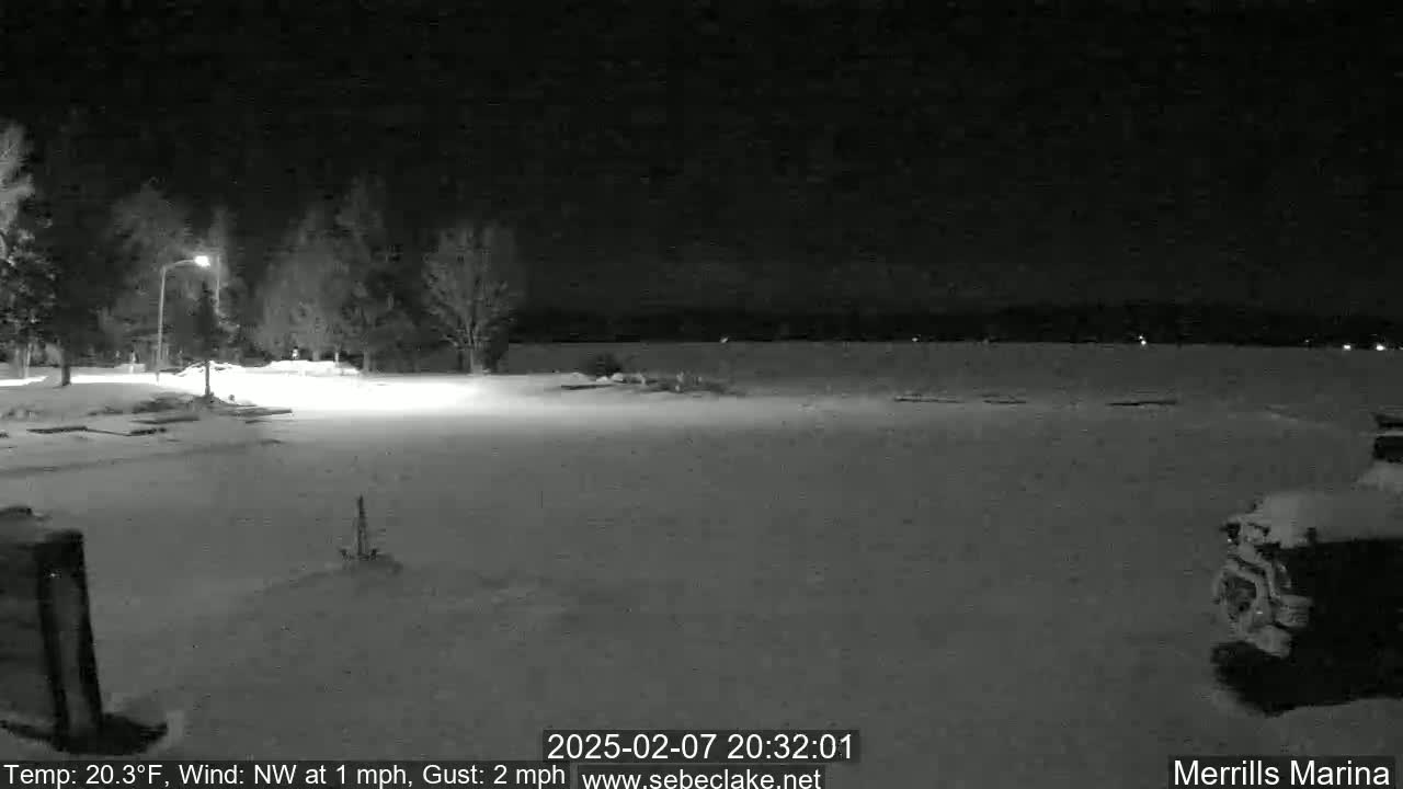 A snow-covered nighttime parking lot next to a dark body of water under a clear sky with a light northwest wind.