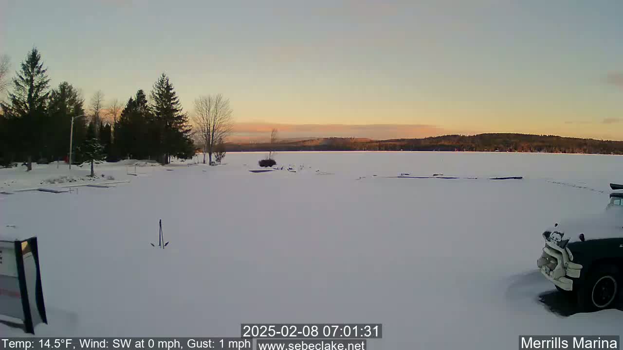 A snow-covered frozen lake at sunrise, with evergreen trees lining the shore and a vehicle parked nearby.