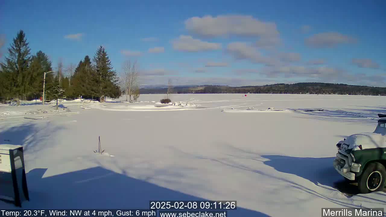 A snow-covered, frozen lake is seen under a mostly sunny sky, with evergreen trees lining the shore and a partially visible truck parked in the snow.