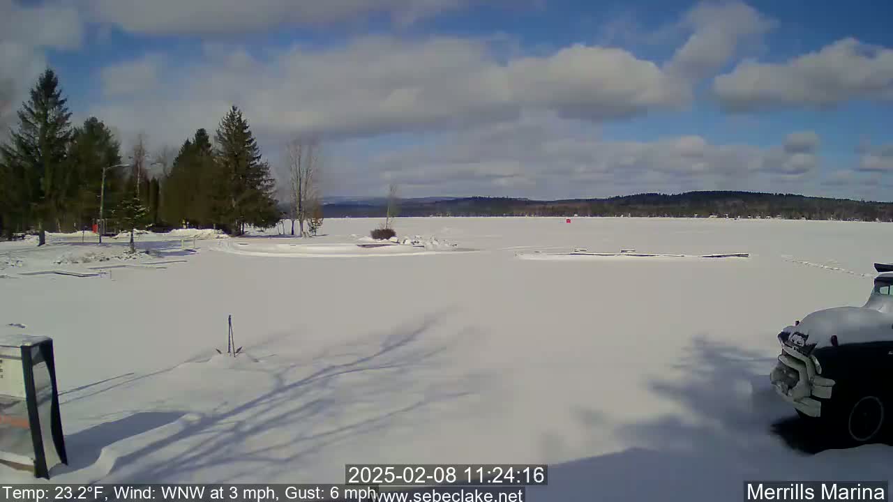 A snow-covered shoreline and frozen lake are visible under a partly cloudy, sunny sky, with a dark-colored truck parked in the lower right corner.