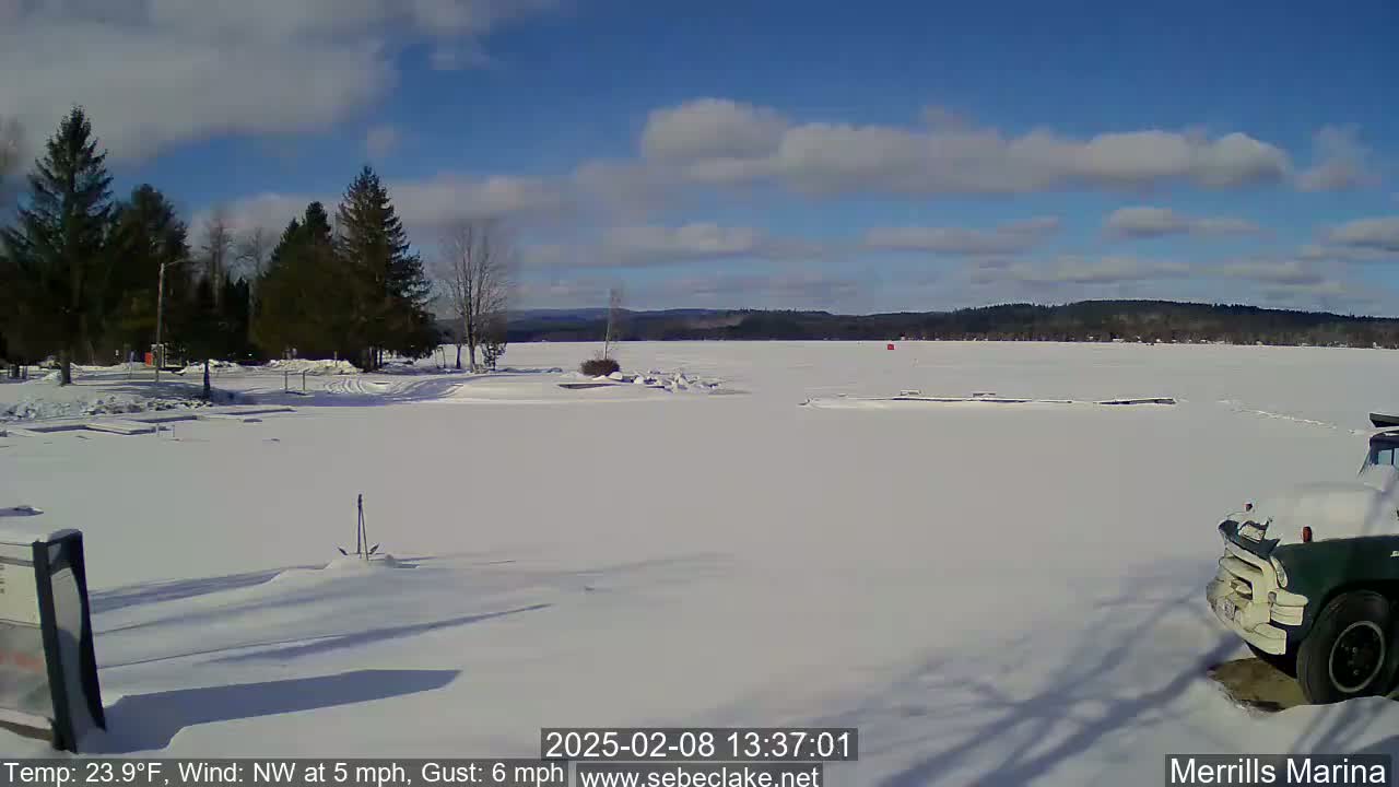A snow-covered frozen lake is seen under a partly cloudy blue sky, with a few evergreen trees and a dark-colored truck partially visible in the foreground.