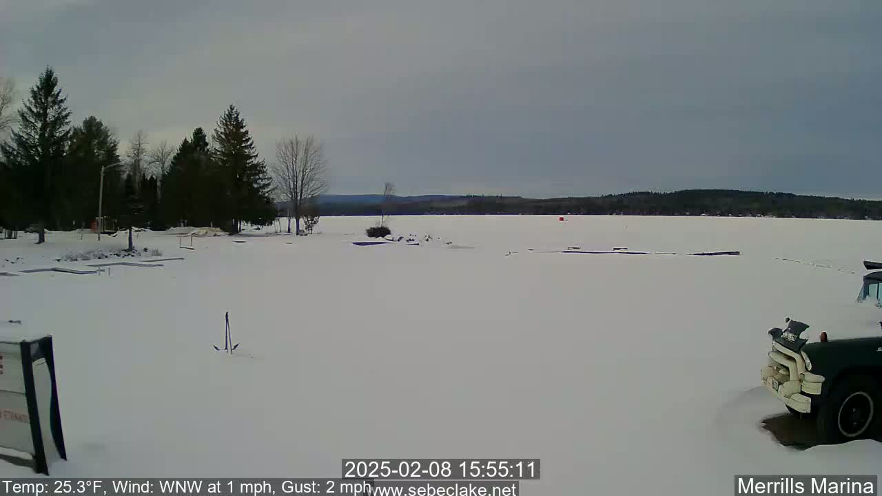 A snow-covered frozen lake is seen beyond a snow-covered parking lot with a few evergreen trees and a dark-colored truck parked in the lower right corner under an overcast sky.