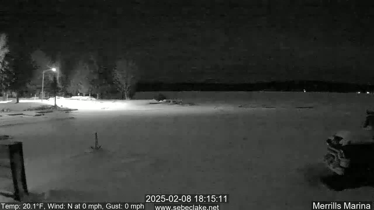 A snow-covered nighttime scene shows a marina area with a body of water in the background under a clear, dark sky.