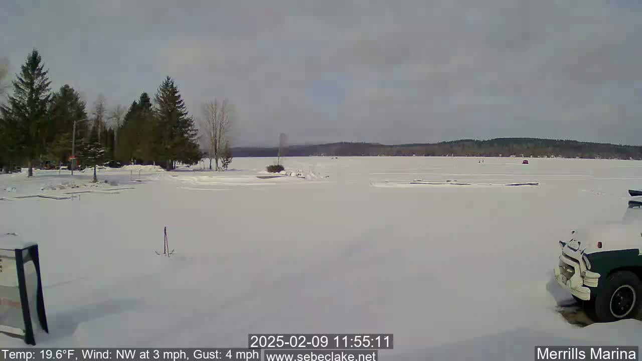 A snow-covered frozen lake is seen from a shoreline with evergreen trees in the background under an overcast sky.