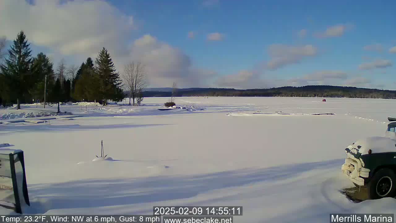 A snow-covered frozen lake is seen under a partly cloudy, sunny sky, with evergreen trees and a partially visible vehicle in the foreground.