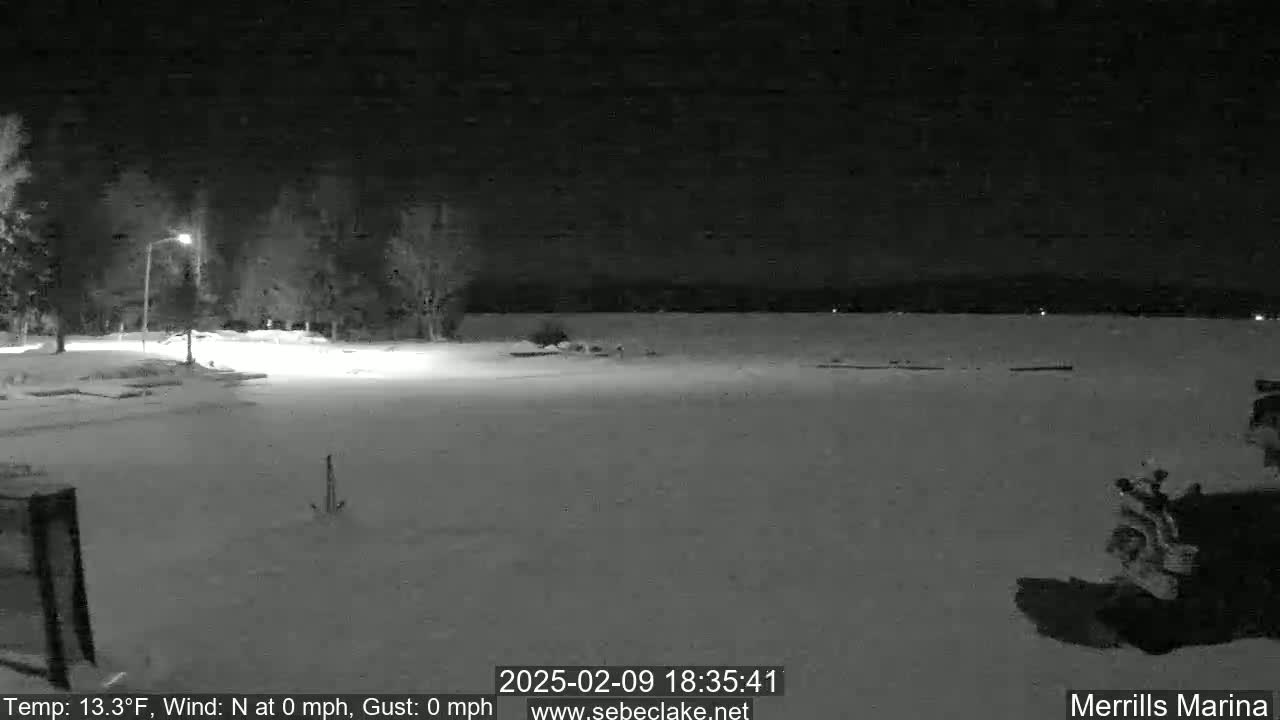 A snow-covered nighttime scene shows a parking lot near a body of water under a clear, dark sky.