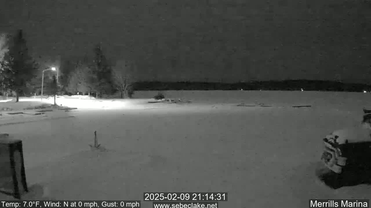 A snow-covered parking lot and marina are visible at night under a dark sky, with a few trees and lights in the background.