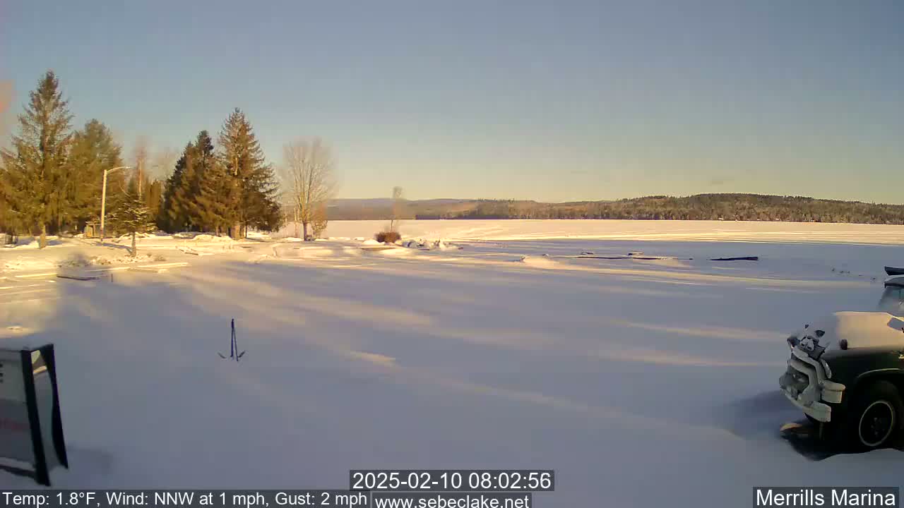 A snow-covered parking lot and frozen lake are bathed in the morning sun under a clear sky.