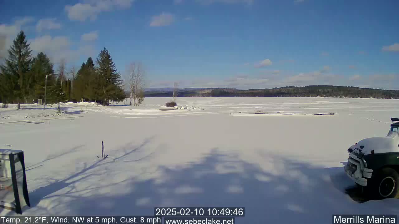 A snow-covered frozen lake is seen under a clear blue sky, with evergreen trees lining the shore in the foreground and a partially visible vehicle parked near the lower-right corner.