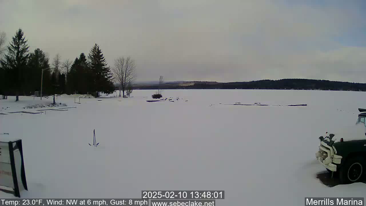 A snow-covered frozen lake is seen from a shoreline with evergreen trees, a partially visible vehicle, and a mostly cloudy sky.