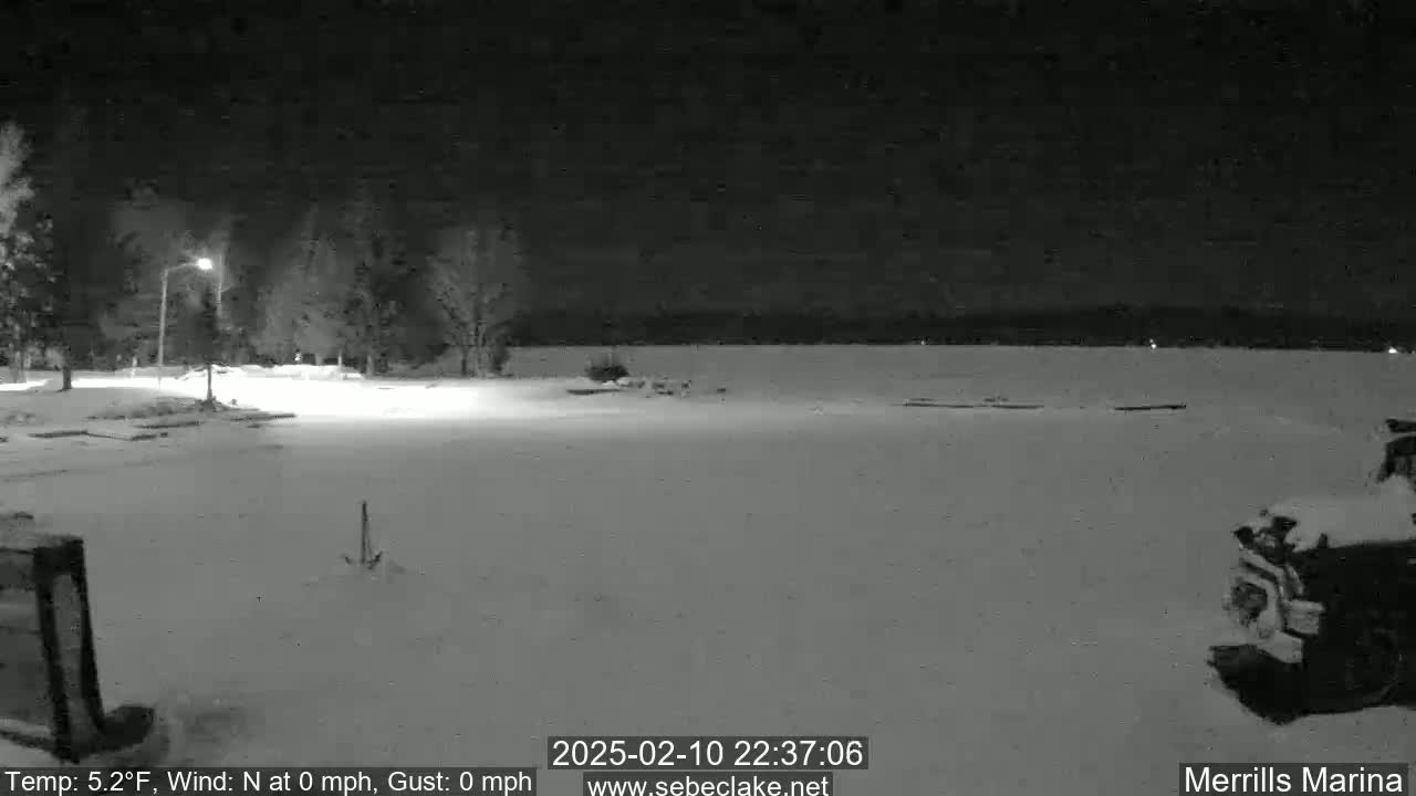 A snow-covered nighttime scene shows a body of water in the distance, a few trees, and a snowplow partially visible in the bottom right corner.