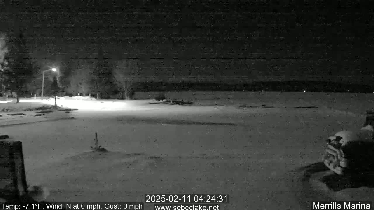 A snow-covered nighttime scene shows a mostly empty parking lot next to a body of water under a dark sky.