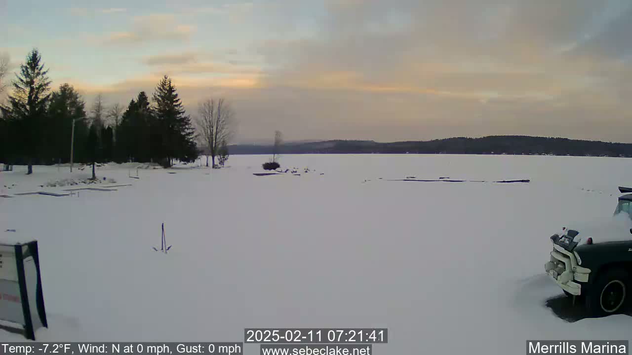 A snow-covered frozen lake is visible under a pale sunset sky, with a few dark evergreen trees and a partially visible dark truck in the foreground.