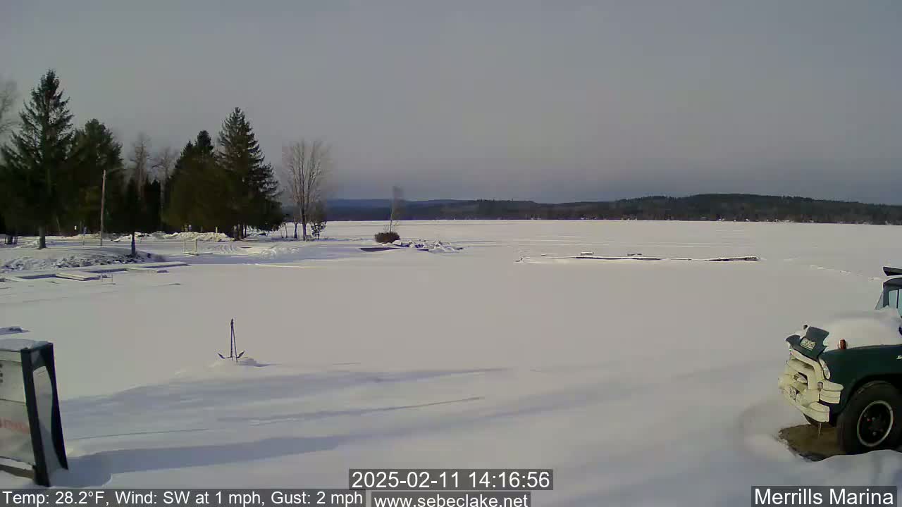 A snow-covered frozen lake is seen from a shoreline with evergreen trees and a partially visible truck in the lower right corner under a clear, bright sky.