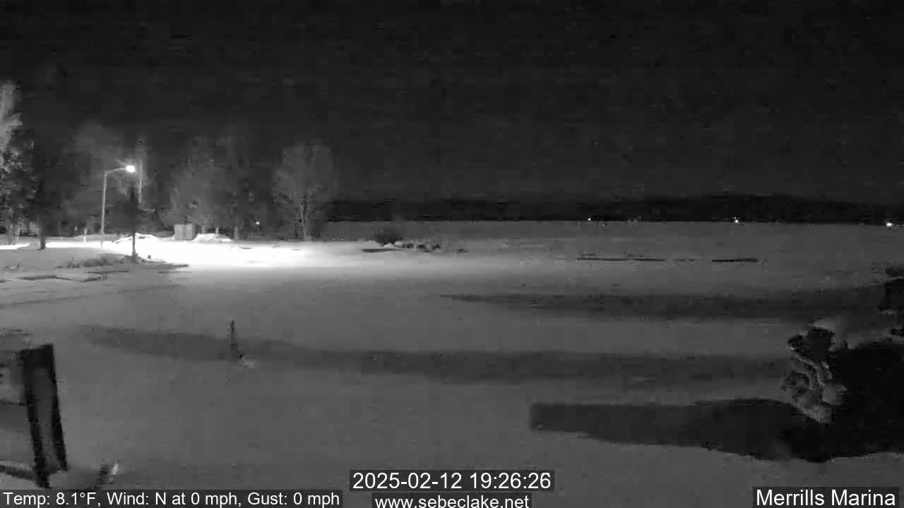 A nighttime, snow-covered scene shows a mostly empty parking lot near a body of water under a clear, dark sky.