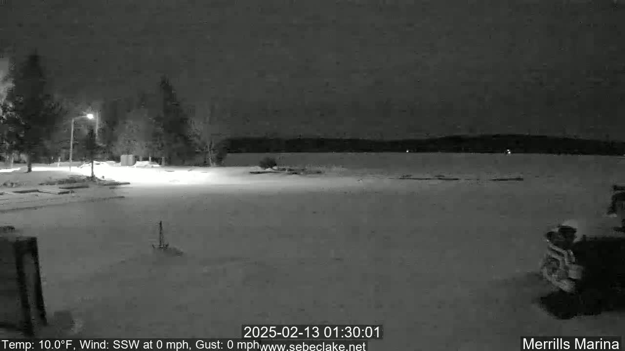 A snow-covered nighttime scene shows a frozen lake, a few trees, and a vehicle in the foreground, under a dark sky.