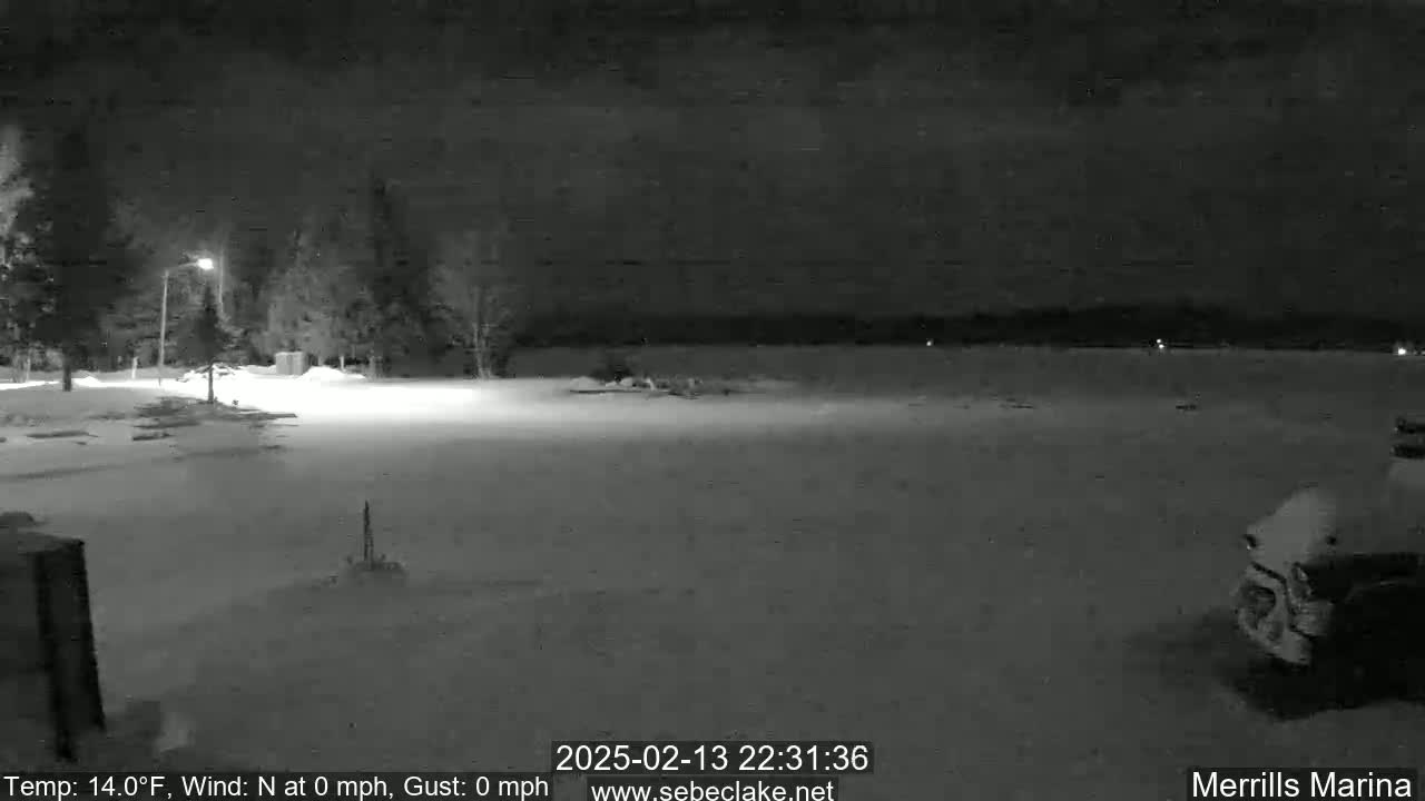 A snow-covered nighttime scene shows a mostly empty parking lot near some trees and a body of water under a dark, clear sky.