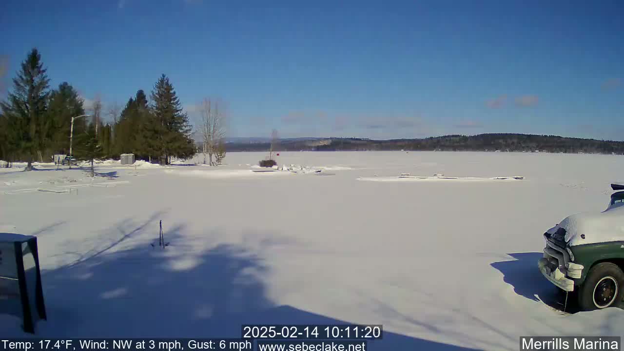 A snow-covered frozen lake is seen under a clear blue sky, with evergreen trees in the foreground and a partially visible truck parked in the snow.