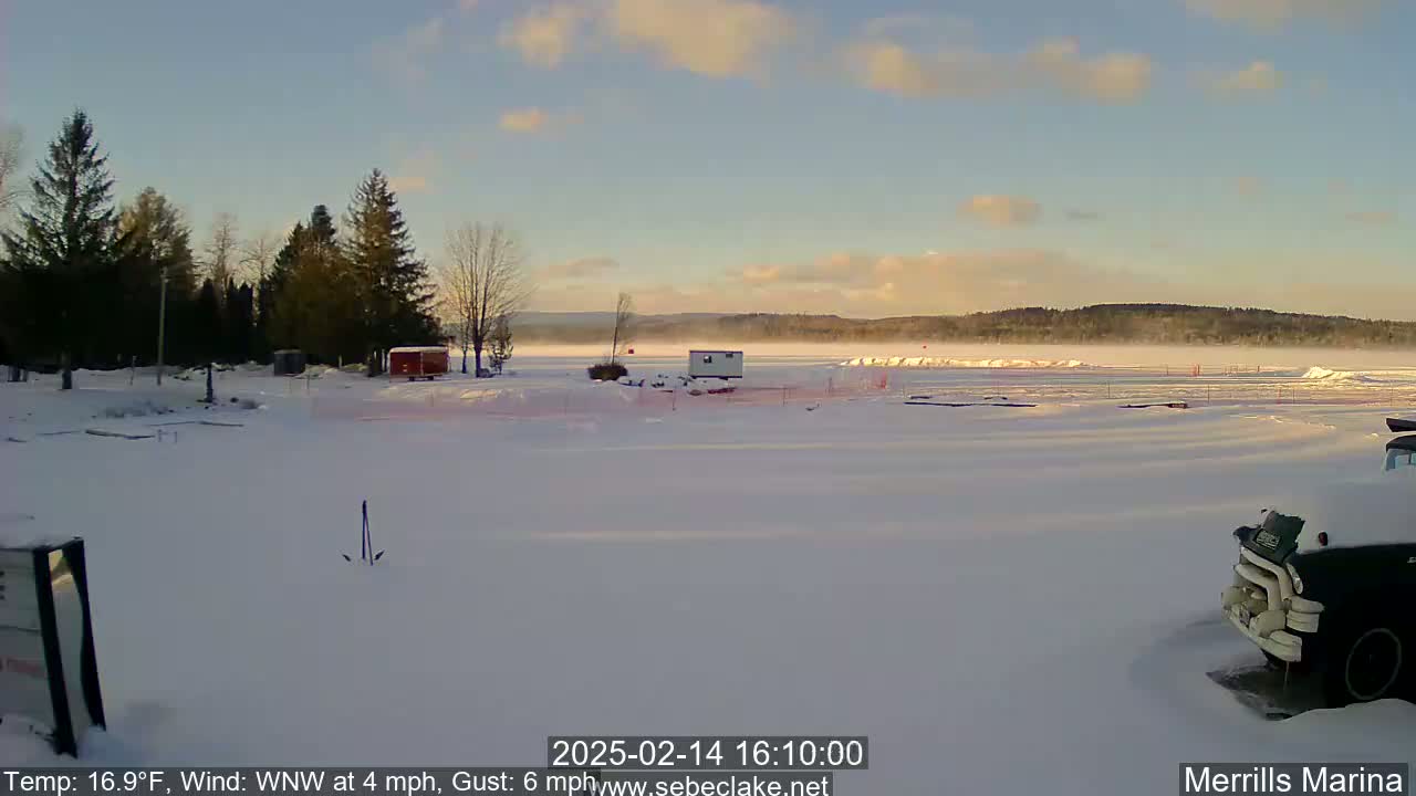 A snow-covered marina with a frozen lake in the background under a mostly clear, light blue sky.