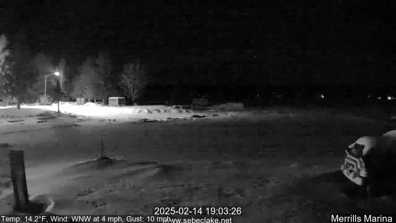 A snow-covered nighttime scene shows a mostly empty parking lot near some trees and a building, with a vehicle parked in the lower right corner under a dark, possibly windy sky.