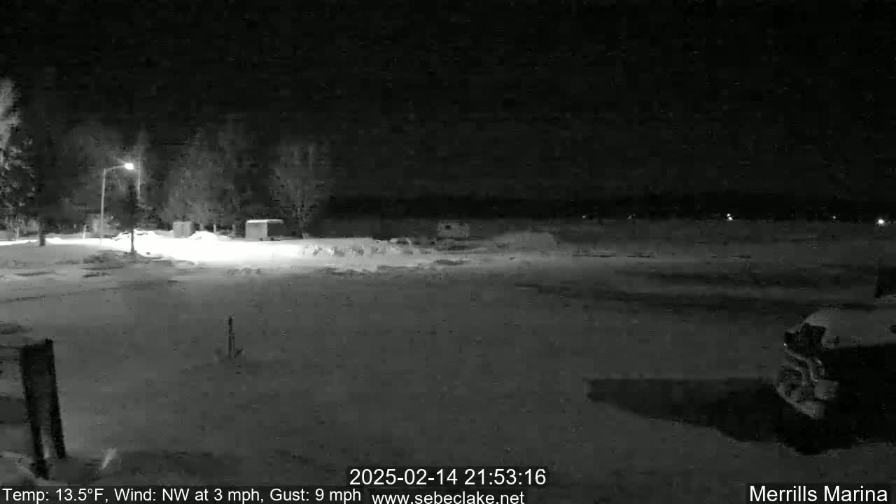 A snow-covered nighttime scene shows a sparsely populated parking lot with a few vehicles and trees under a dark sky.