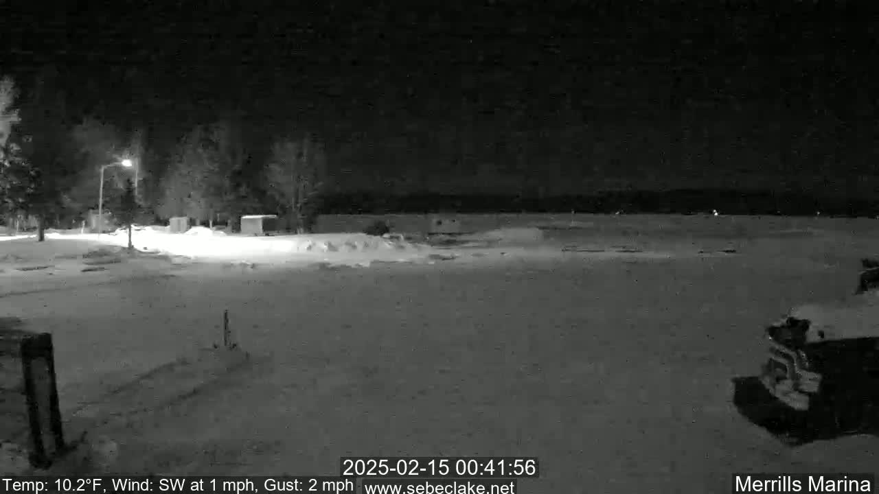 A snow-covered nighttime scene at a marina shows a vehicle parked near a sparsely lit area with trees in the background under a dark sky.
