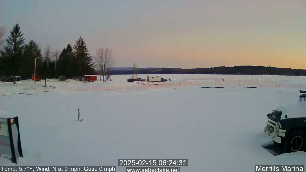 A snow-covered parking lot near a frozen lake is shown at dawn, with a few vehicles visible and trees in the background.