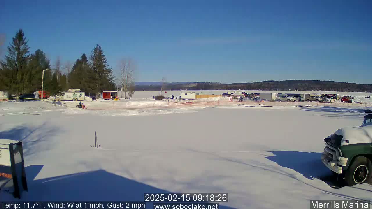 A snow-covered parking lot and frozen lake are visible under a clear, sunny sky, with several vehicles and people present.