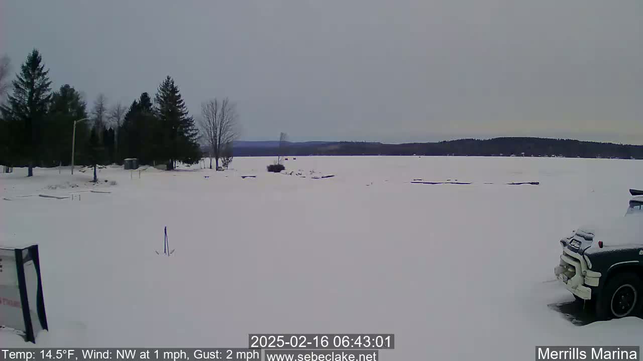 A snow-covered frozen lake is seen from a shoreline with a few evergreen trees, a parked truck, and overcast skies.