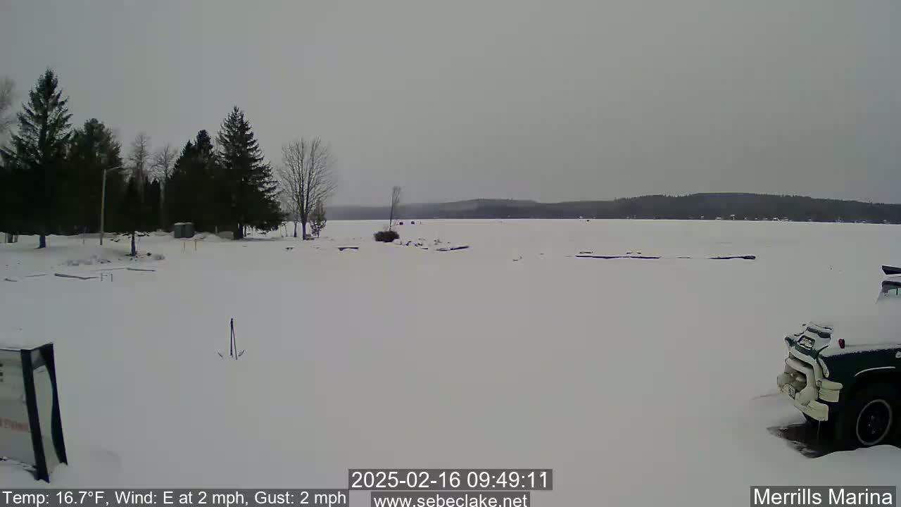 A snow-covered, frozen lake is seen from a shoreline with dark evergreen trees, a partially visible vehicle, and overcast skies.