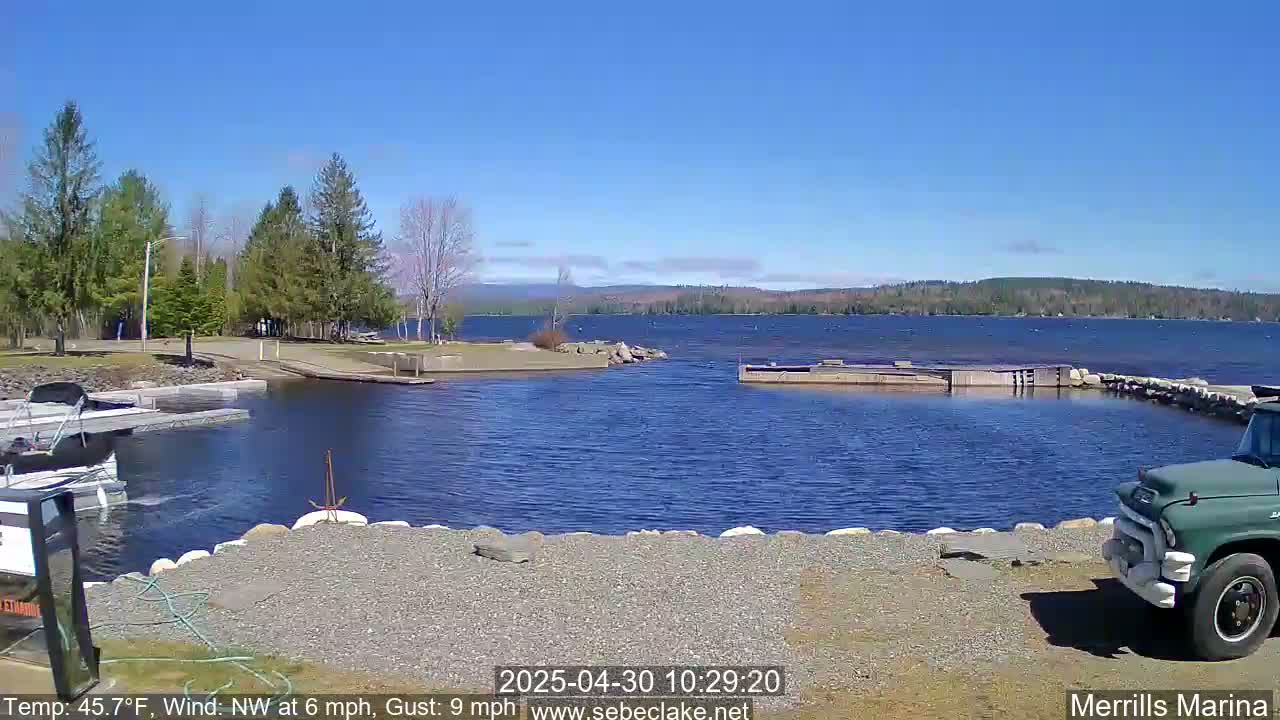 A sunny day at a marina shows a calm lake with a shoreline of trees, a gravel area, some docked boats, and a green truck.