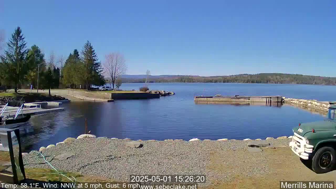 A calm, sunny day at a marina shows a tranquil lake with a rocky shoreline, boats docked at a pier, and a green truck parked nearby.