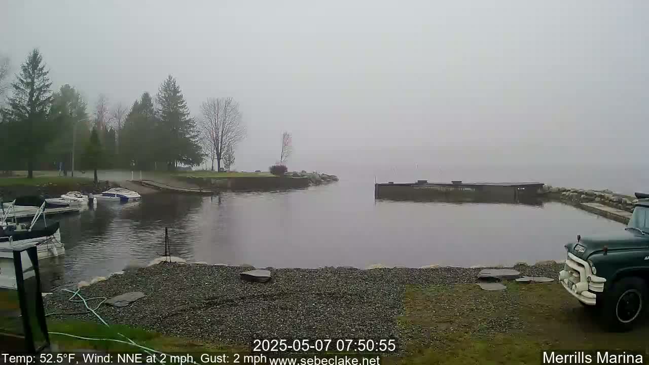 A foggy marina scene shows several boats moored near a rocky shoreline, with a dark-colored truck parked nearby.