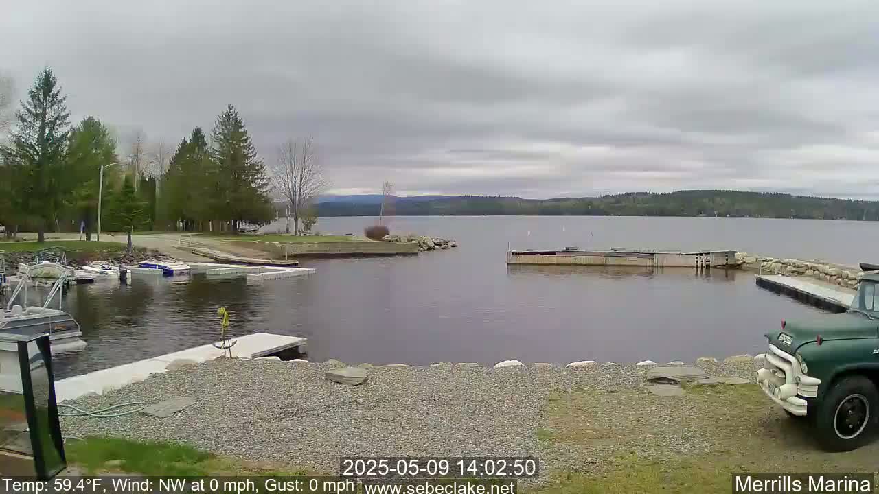 A calm lake with overcast skies, a gravel shoreline, several boats docked, and a green truck parked near a marina.