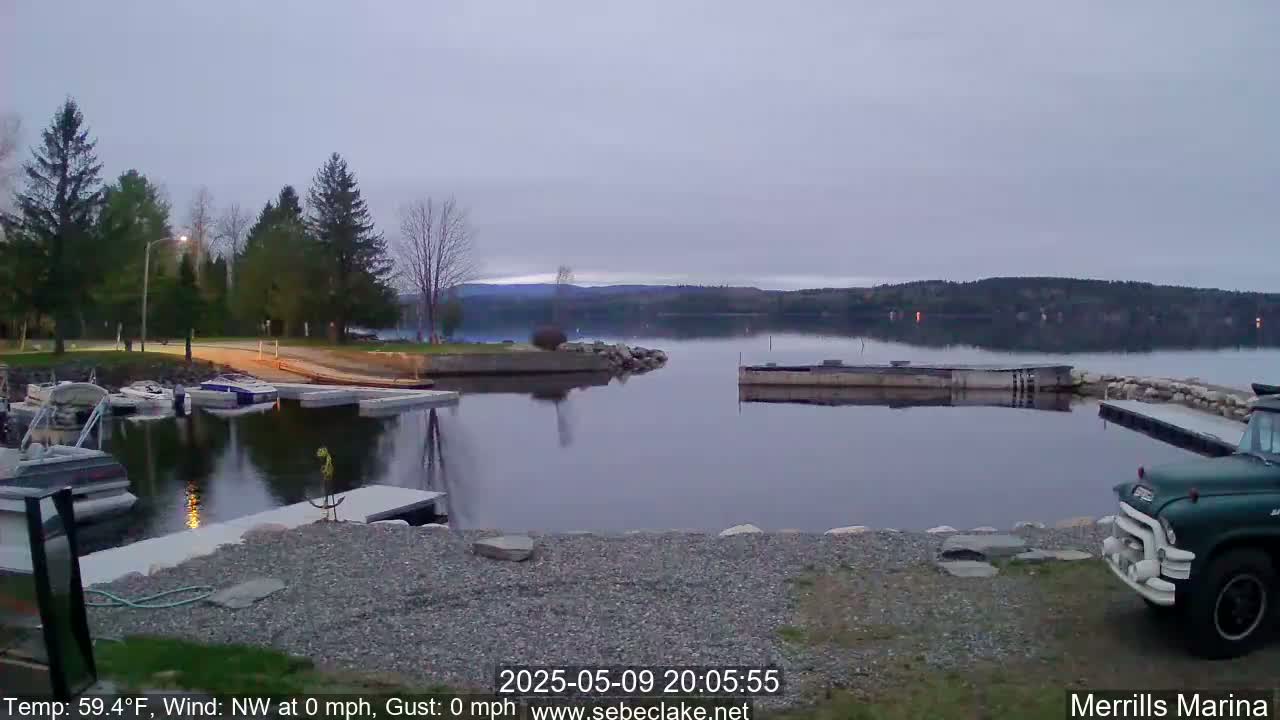 A calm, twilight scene shows a marina with boats docked, a gravel shoreline, and a lake extending to a dark, wooded hillside under a pale, overcast sky.