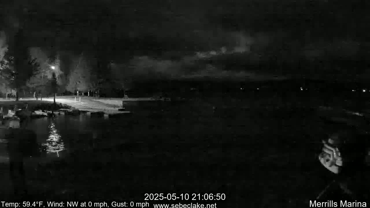 A nighttime, black and white view of a calm body of water next to a dimly lit shoreline under a dark, cloudy sky.