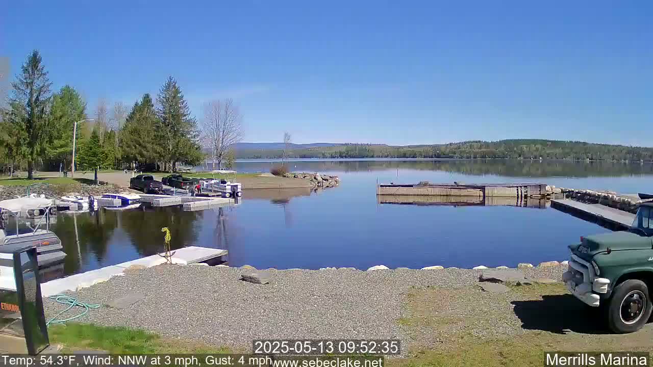 A sunny day at a marina shows calm water, boats docked, and a truck parked on a gravel shore, with trees and a distant shoreline visible.