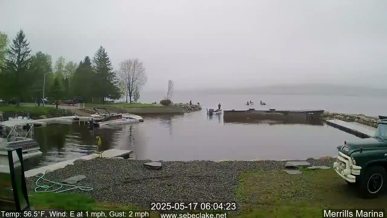A foggy marina scene shows several boats docked and some people fishing on a calm lake, with green trees lining the shore.