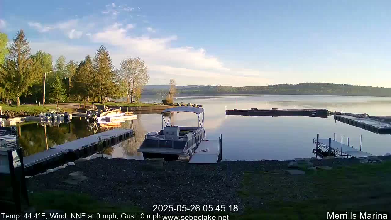 A calm lake at sunrise, under a partly cloudy sky, is shown with several boats docked at a marina surrounded by trees.