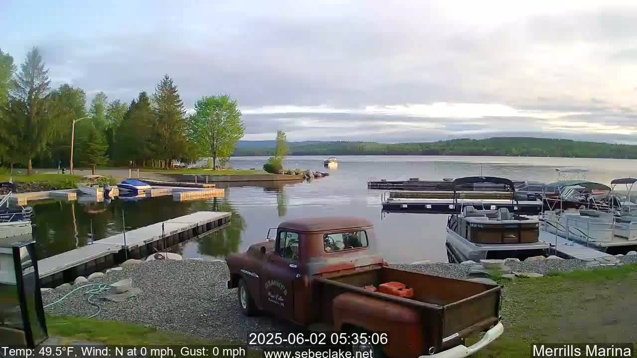 A rusty pickup truck is parked at a marina on a calm lake under an overcast sky, with several boats docked nearby and trees lining the shore.