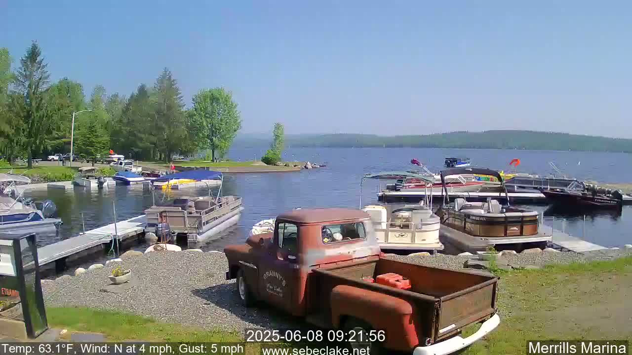 A rusty red pickup truck is parked at a marina on a sunny day with a light north wind, surrounded by numerous pontoon boats and other watercraft docked at the water's edge.