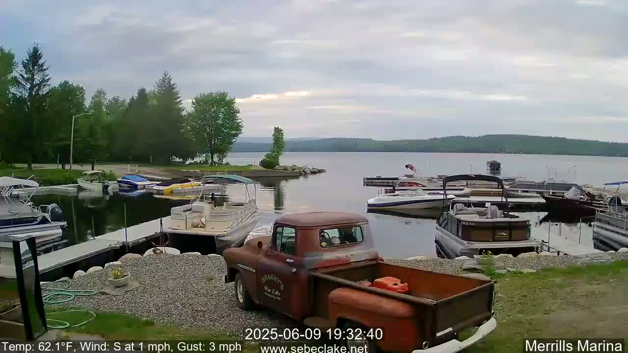 A rusty red pickup truck sits in a marina with numerous boats docked along a calm lake under an overcast sky.