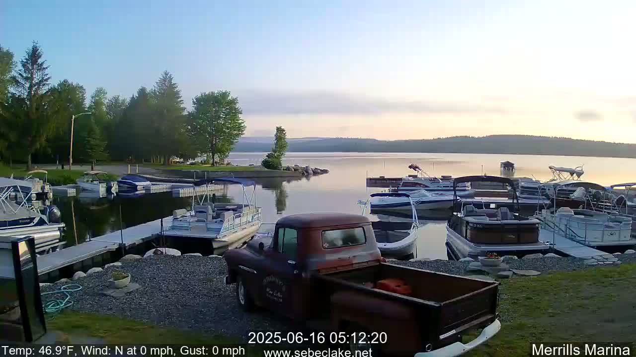 A calm, sunrise illuminates a marina filled with numerous pontoon boats and other watercraft, with a rusty pickup truck parked nearby.