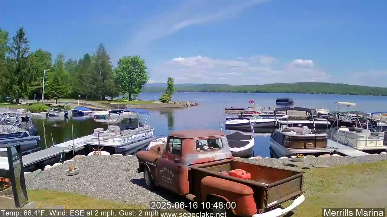 A rusty red pickup truck sits in a marina on a sunny day with a calm lake in the background, surrounded by numerous docked boats and lush green trees.