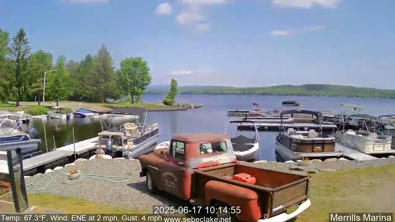A rusty red pickup truck sits in a marina on a sunny day with a calm lake in the background, numerous boats docked along the shoreline and green trees lining the water's edge.