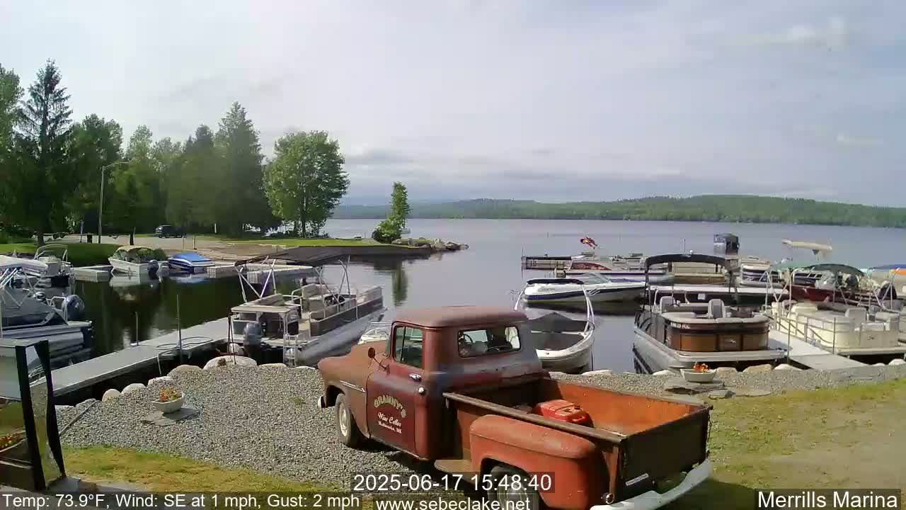A rusty red pickup truck sits in a marina next to a calm lake surrounded by green trees on a partly sunny day with a southeasterly wind.