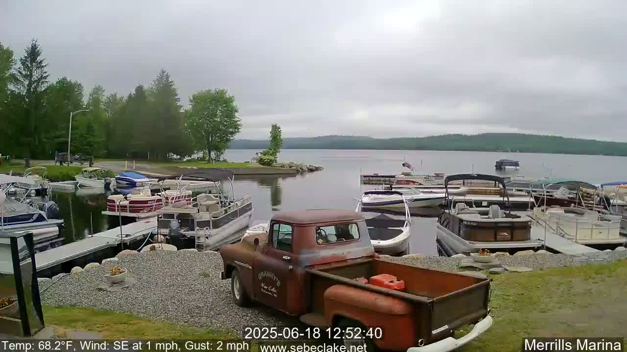 A rusty red pickup truck sits in a marina filled with pontoon boats on a cloudy day.