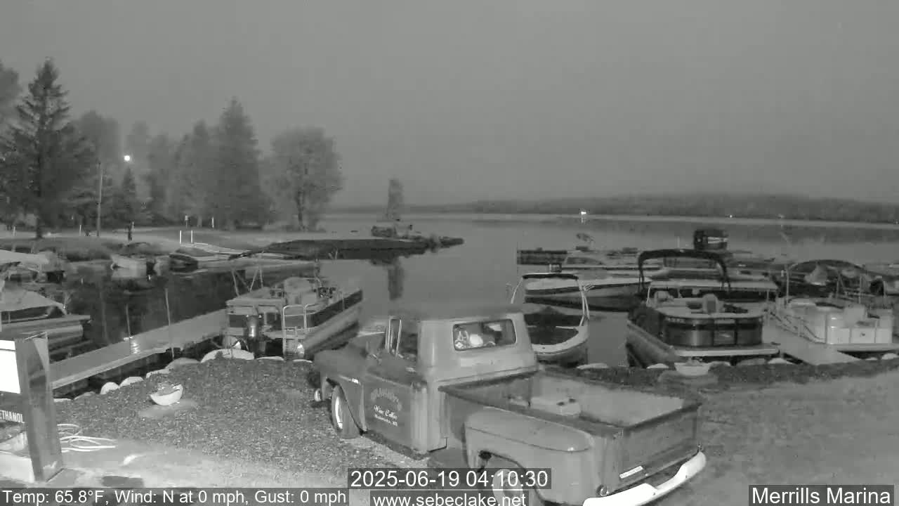 A nighttime view of a marina with numerous boats docked, a pickup truck, and calm water under hazy conditions.